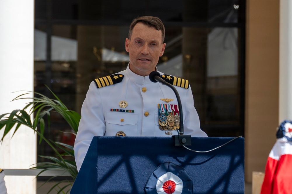 A senior naval officer in white dress uniform is speaking at a podium during what appears to be a formal military ceremony. The officer displays multiple medals, ribbons, and insignia consistent with a high-ranking U.S. Navy Captain. An American flag is partially visible in the background to the right.