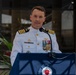 A senior naval officer in white dress uniform is speaking at a podium during what appears to be a formal military ceremony. The officer displays multiple medals, ribbons, and insignia consistent with a high-ranking U.S. Navy Captain. An American flag is partially visible in the background to the right.