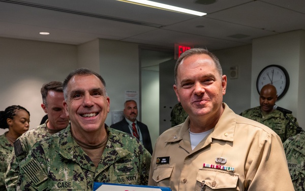 Two U.S. Navy officers are posing together while shaking hands and jointly holding a Legion of Merit certificate. The officer on the right is wearing the Legion of Merit medal on his khaki uniform, indicating he is the award recipient. Several other Navy personnel are visible in the background of what appears to be an indoor military facility.