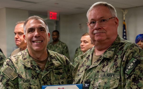 Two U.S. Navy officers are posing together while jointly holding a Meritorious Service Medal certificate. The officer on the left, identified by his name tape as 'CASE,' is presenting the award to the officer on the right, identified as 'SCHMIDT,' who is wearing the medal on his uniform. The ceremony is taking place indoors with other military personnel visible in the background.