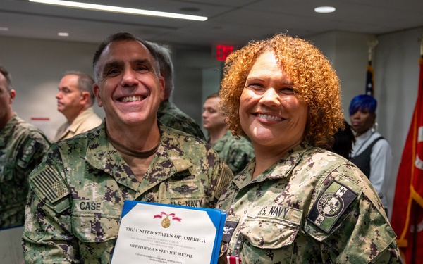 Two U.S. Navy personnel in NWU Type III camouflage uniforms pose together while holding a Meritorious Service Medal certificate. The individual on the left, identified by name tape as 'CASE,' is presenting the award to the individual on the right, who is wearing the medal on their uniform. The ceremony appears to be taking place in an indoor military facility.
