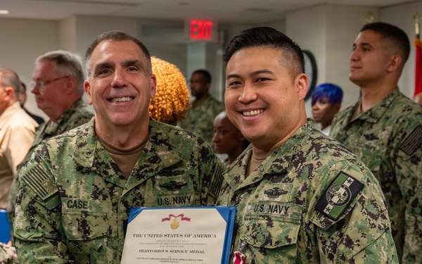 Two U.S. Navy personnel in digital camouflage uniforms (NWU Type III) pose together holding a Meritorious Service Medal certificate. The officer on the left, identified by his name tape as 'CASE,' is presenting the award to the officer on the right, who is wearing the medal on his chest. The ceremony appears to be taking place indoors with other Navy personnel visible in the background.