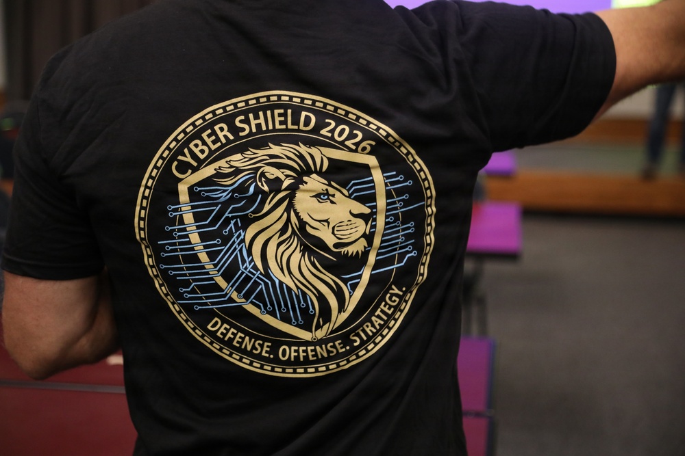 The image shows the back of a person wearing a black t-shirt featuring a large circular graphic design. The design prominently displays 'CYBER SHIELD 2026' at the top and 'DEFENSE. OFFENSE. STRATEGY.' at the bottom, with a gold lion head on a shield surrounded by circuit board patterns. The setting appears to be an indoor venue with blurred background elements.