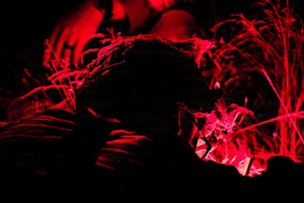 A nighttime photograph taken under red light illumination, showing one or more individuals crouched or lying low among tall grass or reeds. The scene is heavily shadowed with only red-lit outlines visible, creating a dramatic, high-contrast composition. The subject appears to be engaged in a ground-level activity in a natural outdoor setting.