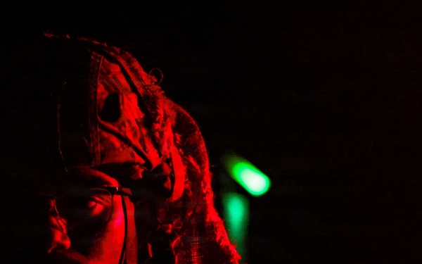 A person wearing tactical military-style headgear and protective eyewear is illuminated by a red light source against a near-total black background. A small green light is visible in the background, consistent with a chemical light stick or indicator light. The dramatic red lighting and dark environment suggest a nighttime or low-light operational setting.