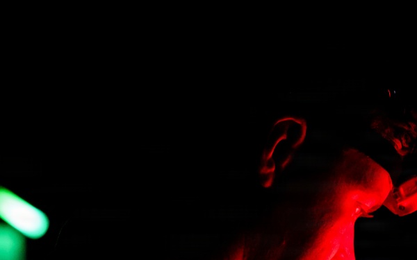 A person is photographed in profile view against a near-total black background, illuminated dramatically by a red light source from the right side. A distinct green light source is visible in the background to the left, creating a strong color contrast. The image has a dark, atmospheric quality consistent with low-light documentary or artistic photography.