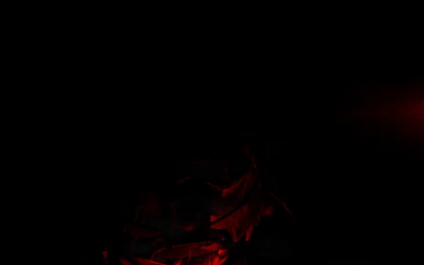 A military personnel is examining what appears to be a topographic map in complete darkness, illuminated solely by a red-light headlamp. The red light is used deliberately to preserve night vision while allowing map reading. The subject is wearing camouflage uniform, a face covering, and protective eyewear.