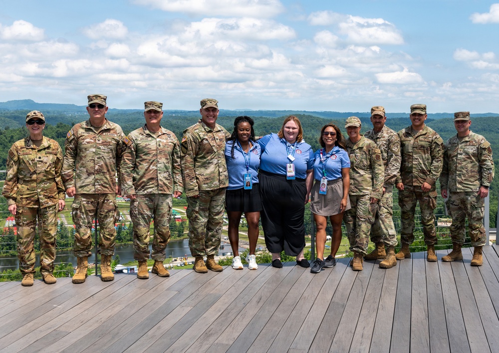 A group of approximately eleven individuals pose for a formal group photo on an elevated outdoor deck or observation platform. The group consists of eight military personnel in U.S. Army OCP (Operational Camouflage Pattern) uniforms and three civilians wearing matching light blue polo shirts with lanyards and ID badges. The backdrop features a scenic landscape with forested hills, a river or lake, and a partially cloudy sky.