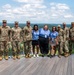 A group of approximately eleven individuals pose for a formal group photo on an elevated outdoor deck or observation platform. The group consists of eight military personnel in U.S. Army OCP (Operational Camouflage Pattern) uniforms and three civilians wearing matching light blue polo shirts with lanyards and ID badges. The backdrop features a scenic landscape with forested hills, a river or lake, and a partially cloudy sky.
