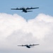 Two military turboprop transport aircraft are flying in formation against a partly cloudy sky. Both aircraft appear to be C-130 Hercules or similar variants, painted in gray military livery. The upper aircraft is closer to the camera and more detailed, while the second flies at a lower altitude in the background.