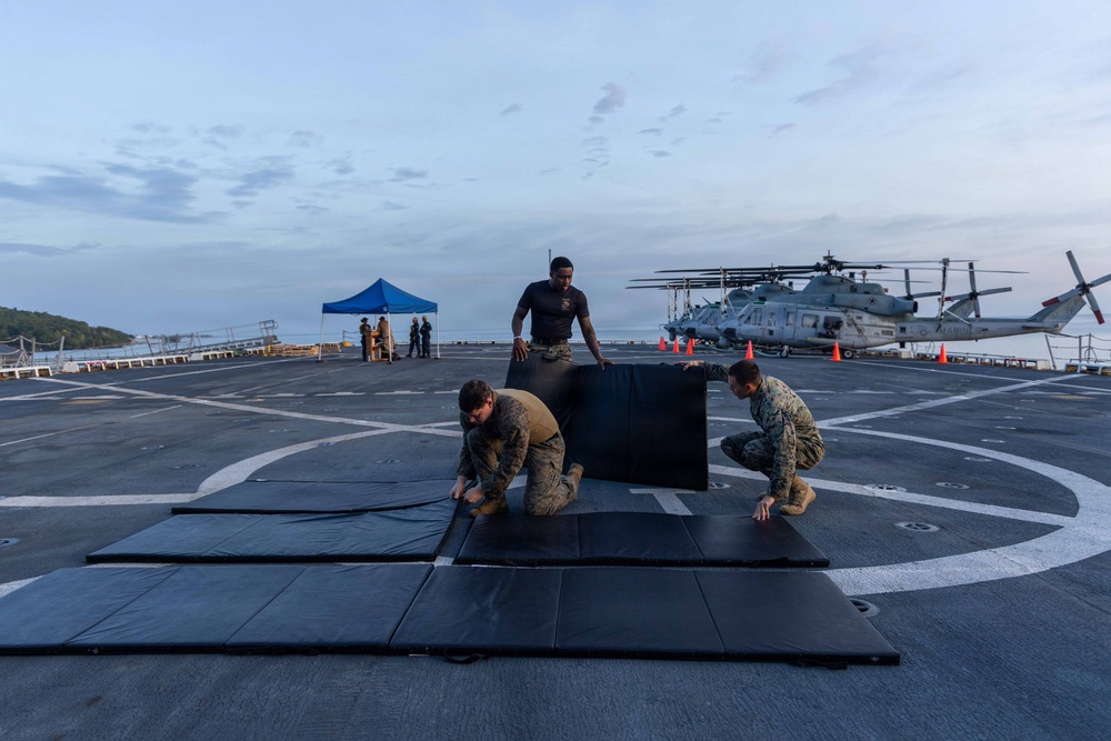 Three military personnel in camouflage and dark uniforms are setting up black exercise/training mats on the flight deck of a naval vessel. The scene appears to be at dawn or dusk, with helicopters visible in the background along with orange safety cones and a blue canopy tent. The activity suggests preparation for a physical training or martial arts session on the ship's deck.