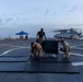 Three military personnel in camouflage and dark uniforms are setting up black exercise/training mats on the flight deck of a naval vessel. The scene appears to be at dawn or dusk, with helicopters visible in the background along with orange safety cones and a blue canopy tent. The activity suggests preparation for a physical training or martial arts session on the ship's deck.