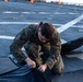 A U.S. Marine in woodland MARPAT camouflage uniform is kneeling on what appears to be a ship's flight deck, working with a large black mat or tarp. The marine appears to be folding, unfolding, or securing the black material on the deck surface. Orange traffic cones and painted deck markings are visible in the background, consistent with a naval vessel flight deck environment.
