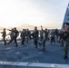 A group of military personnel, predominantly U.S. Marines in MARPAT camouflage uniforms, are conducting martial arts or physical training drills on the flight deck of a naval vessel. The training appears to involve kicking and striking techniques, consistent with Marine Corps Martial Arts Program (MCMAP) exercises. The scene is set at dawn or dusk with a coastal waterway visible in the background.