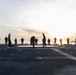 A group of military personnel are gathered on the flight deck of a naval vessel during sunrise or sunset. The silhouetted figures appear to be engaged in physical training or a formation activity on the ship's deck. The low-angle sun creates strong backlighting, casting the personnel and ship structures in silhouette against a golden horizon over open water.