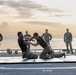 Two military personnel in black shirts and camouflage trousers are engaged in a hand-to-hand combat training exercise on padded mats aboard what appears to be a naval vessel's deck. Several uniformed observers, wearing full camouflage uniforms consistent with U.S. Marine Corps MARPAT pattern, stand in a semicircle watching the demonstration. The scene is set at dusk or dawn with a calm ocean and cloudy sky visible in the background.