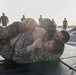 Two military personnel in camouflage uniforms are engaged in a ground combat/grappling training exercise on black mats. The training appears to take place on a ship's deck or military installation, with multiple uniformed observers watching in the background. The scene depicts what appears to be Marine Corps Martial Arts Program (MCMAP) or similar hand-to-hand combat training.