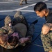 Two individuals in U.S. Marine Corps MARPAT camouflage uniforms are engaged in a ground grappling drill on black training mats. A third person in a black t-shirt and camouflage shorts observes closely, likely acting as an instructor or referee. The setting appears to be an outdoor hard surface, possibly a ship deck or tarmac, with additional personnel visible in the background.