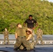 A group of military personnel, appearing to be U.S. Marines based on their distinctive MARPAT camouflage uniforms, are conducting a martial arts or combatives training session on what appears to be a ship's deck or elevated platform. A central instructor in a black t-shirt with a Marine Corps emblem oversees two trainees grappling on a mat. Additional Marines observe in the background against a lush, forested hillside.