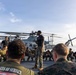 A military instructor or non-commissioned officer stands and addresses a group of Marines gathered in a circle on the flight deck of a naval vessel. The personnel are wearing camouflage uniforms and physical training attire, crouching or squatting around the standing figure. Military helicopters are parked on the flight deck in the background, with open ocean visible on the horizon.