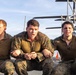 Three male individuals wearing MARPAT (Marine pattern) camouflage uniforms are seated together on what appears to be a ship's flight deck, with a helicopter visible in the background. They appear to be engaged in a physical activity or exercise, with expressions suggesting exertion or chanting. The setting and uniforms are consistent with U.S. Marine Corps personnel aboard a naval vessel.