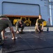 Three military personnel in U.S. Marine Corps uniforms are cleaning or wiping down black exercise/gymnastics mats on what appears to be the deck of a naval vessel. They are working on hands and knees, using white cloths or rags to scrub the mat surface. In the background, a large hangar bay door is visible with yellow heavy equipment (forklift/loader) parked inside.