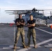 Two U.S. Marines in digital camouflage uniforms and black t-shirts stand with arms crossed on the flight deck of a naval vessel. Behind them is a U.S. Marines helicopter, identifiable by the 'MARINES' marking on its fuselage, with rotor blades folded for deck storage. The setting is an open ocean environment with calm seas visible in the background.
