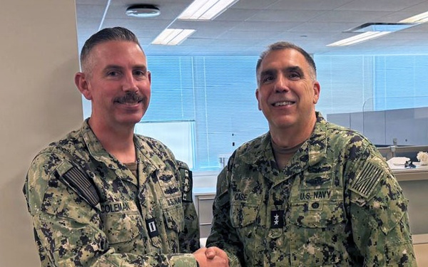 Two U.S. Navy personnel in Navy Working Uniform (NWU Type III) are shaking hands in what appears to be an office environment. The name tapes visible read 'COLEMAN' on the left and 'CASE' on the right, with 'CASE' displaying two stars indicating the rank of Rear Admiral (Upper Half). The setting appears to be a formal recognition or greeting moment within a military administrative facility.