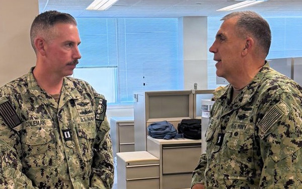 Two U.S. Navy personnel in Navy Working Uniform (NWU Type III) are engaged in conversation inside what appears to be an office environment. The individual on the left is identified by a name tape reading 'COLEMAN' and displays captain's insignia (two silver bars) on the collar. Both wear standard U.S. Navy digital camouflage uniforms with American flag patches on the left shoulder.