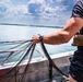 A muscular person standing on a boat is gripping a bundle of dark cables or lines with one hand while wearing a sport watch. The scene is set on open water under a partly cloudy sky. The individual appears to be managing or deploying equipment from the vessel.