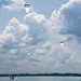 Three parachutists are descending over a large body of water under white and gray round parachutes. A small inflatable boat with personnel is visible on the water surface below, likely serving as a recovery vessel. The scene appears to be a military or training water jump exercise conducted under partly cloudy skies.