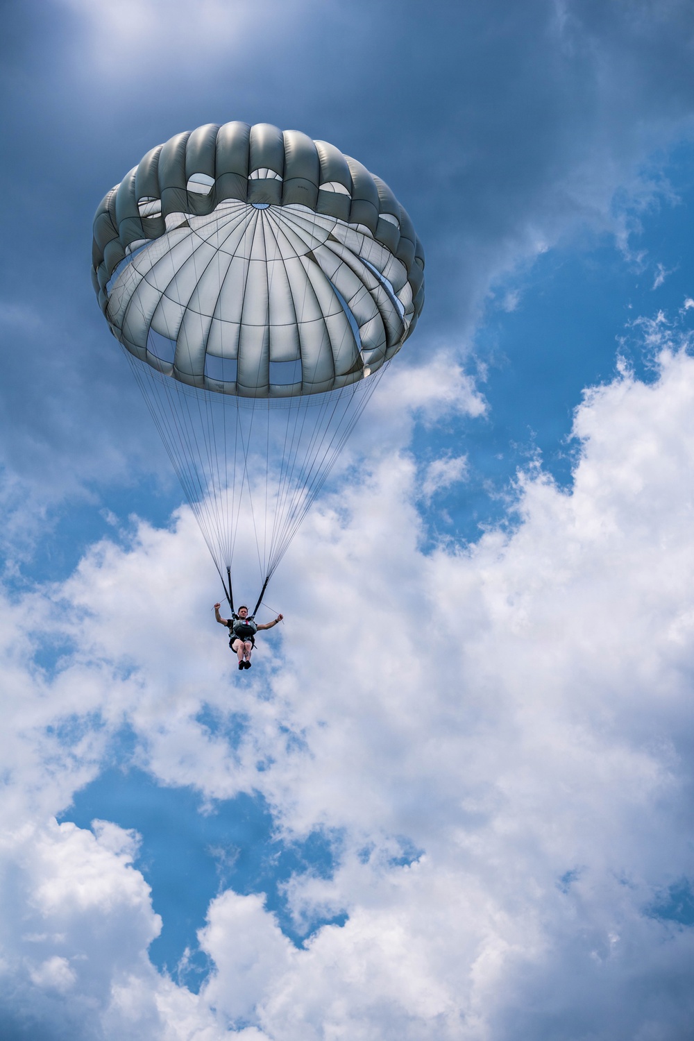 A solo parachutist is descending under a fully deployed round parachute canopy against a partly cloudy blue sky. The canopy is silver-gray in color with visible vents at the top, consistent with a military-style round parachute. The photo is taken from a low angle looking upward, giving a dramatic perspective of the jumper suspended by suspension lines.