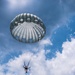A solo parachutist is descending under a fully deployed round parachute canopy against a partly cloudy blue sky. The canopy is silver-gray in color with visible vents at the top, consistent with a military-style round parachute. The photo is taken from a low angle looking upward, giving a dramatic perspective of the jumper suspended by suspension lines.