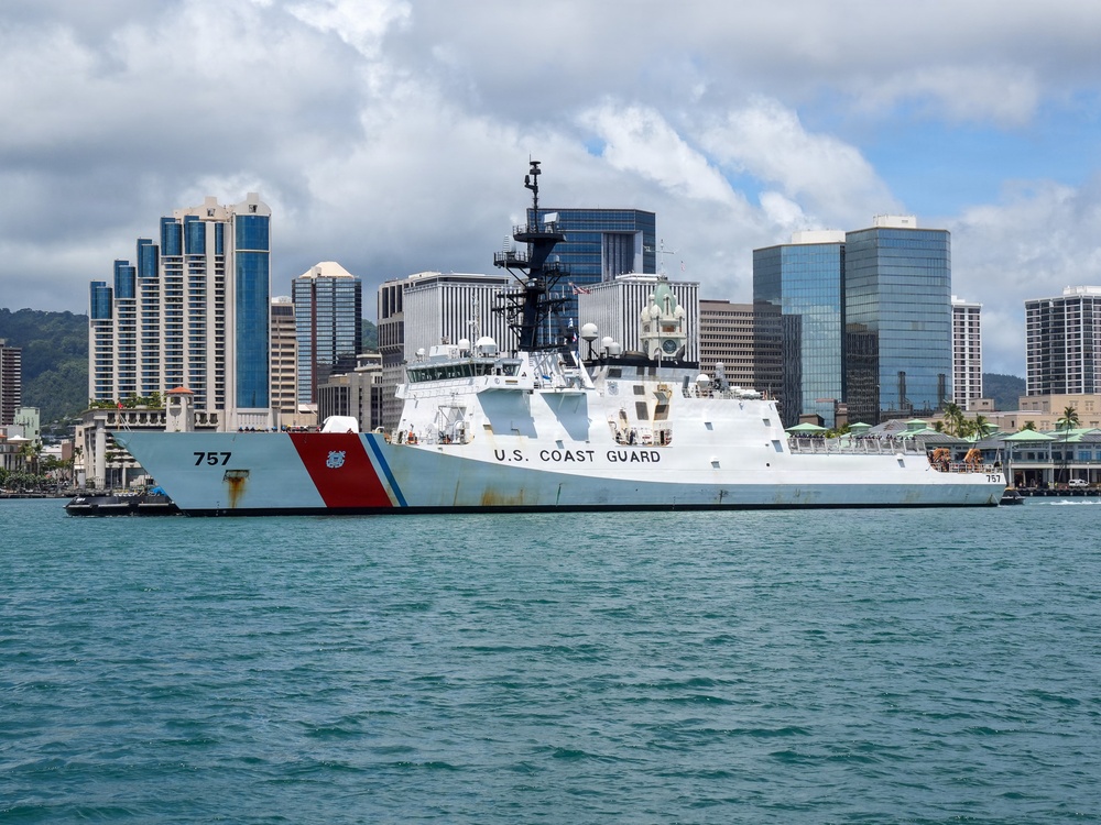 A U.S. Coast Guard cutter bearing hull number 757 is docked at a pier in what appears to be Honolulu Harbor, Hawaii, identifiable by the distinctive downtown skyline in the background. The vessel displays the classic Coast Guard livery with a white hull, red diagonal stripe, and the Coast Guard racing stripe emblem on the bow. The scene is captured in clear daylight with partly cloudy skies over calm turquoise harbor waters.