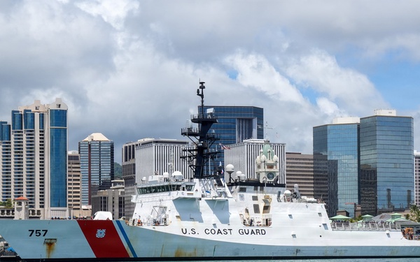 A U.S. Coast Guard cutter bearing hull number 757 is docked at a pier in what appears to be Honolulu Harbor, Hawaii, identifiable by the distinctive downtown skyline in the background. The vessel displays the classic Coast Guard livery with a white hull, red diagonal stripe, and the Coast Guard racing stripe emblem on the bow. The scene is captured in clear daylight with partly cloudy skies over calm turquoise harbor waters.