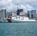 A U.S. Coast Guard cutter bearing hull number 757 is docked at a pier in what appears to be Honolulu Harbor, Hawaii, identifiable by the distinctive downtown skyline in the background. The vessel displays the classic Coast Guard livery with a white hull, red diagonal stripe, and the Coast Guard racing stripe emblem on the bow. The scene is captured in clear daylight with partly cloudy skies over calm turquoise harbor waters.