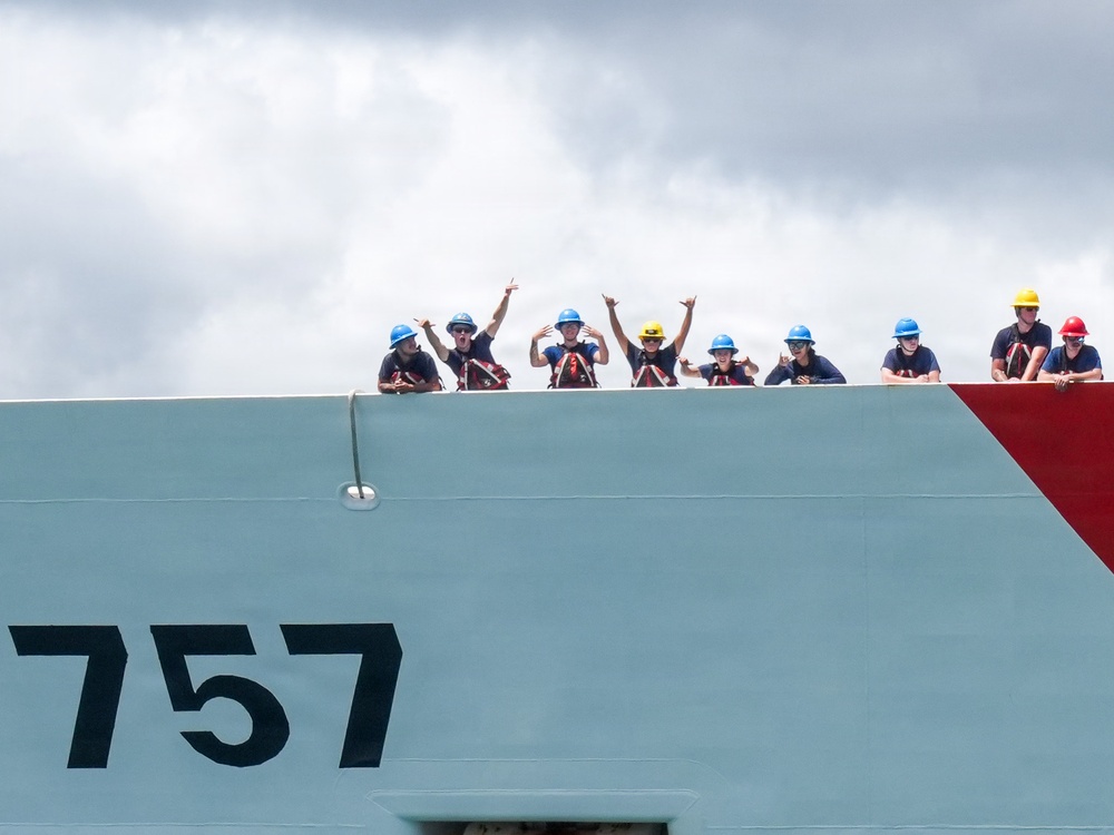 A group of approximately ten crew members are visible standing along the top edge of a large vessel hull marked with the number '757'. The crew members are wearing hard hats in various colors (predominantly blue, with one yellow and one red) and appear to be waving and making hand gestures toward the camera. The vessel features a distinctive red diagonal stripe on its bow, consistent with United States Coast Guard livery.
