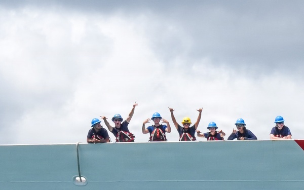 A group of approximately ten crew members are visible standing along the top edge of a large vessel hull marked with the number '757'. The crew members are wearing hard hats in various colors (predominantly blue, with one yellow and one red) and appear to be waving and making hand gestures toward the camera. The vessel features a distinctive red diagonal stripe on its bow, consistent with United States Coast Guard livery.