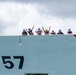 A group of approximately ten crew members are visible standing along the top edge of a large vessel hull marked with the number '757'. The crew members are wearing hard hats in various colors (predominantly blue, with one yellow and one red) and appear to be waving and making hand gestures toward the camera. The vessel features a distinctive red diagonal stripe on its bow, consistent with United States Coast Guard livery.