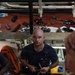 A bald mechanic with a mustache and tattoos on his right arm is working on a large yellow industrial engine, likely in a ship's engine room. He is holding tools and appears to be performing maintenance or repair work. The background shows a workbench covered with numerous scattered tools and mechanical components.