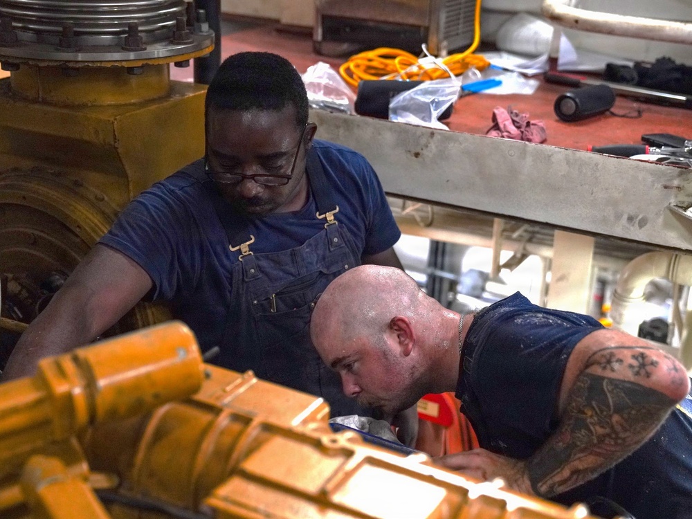 Two workers are performing mechanical maintenance or repair work on large industrial yellow-painted machinery, likely an engine or heavy equipment. One worker wearing glasses and denim overalls observes from above, while a second bald worker with tattoos on his arm leans in closely to inspect or work on a component. The setting appears to be an industrial workspace or engine room with tools and equipment scattered nearby.