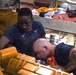 Two workers are performing mechanical maintenance or repair work on large industrial yellow-painted machinery, likely an engine or heavy equipment. One worker wearing glasses and denim overalls observes from above, while a second bald worker with tattoos on his arm leans in closely to inspect or work on a component. The setting appears to be an industrial workspace or engine room with tools and equipment scattered nearby.