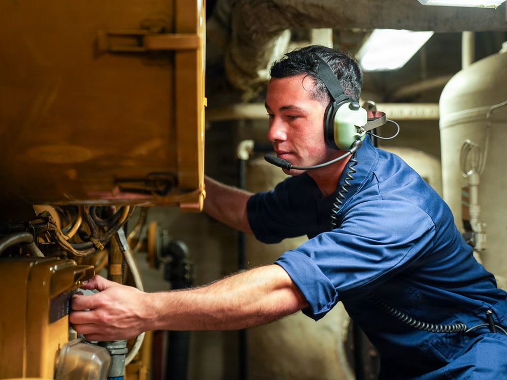 A young male technician wearing a blue uniform and communication headset with microphone is working on industrial or mechanical equipment in a confined, dimly lit space. He is leaning forward and using both hands to manipulate controls or components on a yellow-painted machine or panel. The setting appears to be an engine room, ship compartment, or heavy industrial facility.