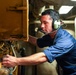 A young male technician wearing a blue uniform and communication headset with microphone is working on industrial or mechanical equipment in a confined, dimly lit space. He is leaning forward and using both hands to manipulate controls or components on a yellow-painted machine or panel. The setting appears to be an engine room, ship compartment, or heavy industrial facility.