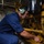 A male technician in blue coveralls is crouching down and performing maintenance on a large yellow industrial engine or machinery system. He is wearing green hearing protection ear muffs, indicating a high-noise environment. The setting appears to be a ship's engine room or mechanical space based on the visible signage and industrial surroundings.
