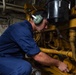 A male technician in blue coveralls is crouching down and performing maintenance on a large yellow industrial engine or machinery system. He is wearing green hearing protection ear muffs, indicating a high-noise environment. The setting appears to be a ship's engine room or mechanical space based on the visible signage and industrial surroundings.