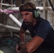 A man wearing green aviation-style headphones with a boom microphone and a black headband is leaning forward in what appears to be an industrial or mechanical workspace. He is wearing a dark blue t-shirt and a black vest, with a visible tattoo on his forearm. The background contains pipes, tubing, and mechanical infrastructure consistent with a ship's interior or large industrial facility.