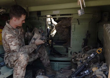 A soldier in desert camouflage uniform is seated inside the rear compartment of a military vehicle, examining or operating a piece of electronic equipment. The vehicle interior contains various military gear including weapons, ammunition boxes, body armor, and communication equipment. The setting appears to be a forward operating environment based on the dusty, worn condition of the equipment.