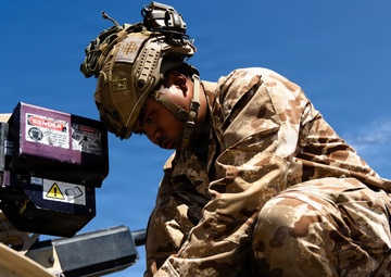A soldier in desert camouflage uniform and tactical helmet is crouched on top of a military vehicle, performing maintenance or technical work. The soldier appears to be connecting or adjusting cables/wiring near a piece of equipment marked with danger warning labels. The image is taken from a low angle against a clear blue sky.