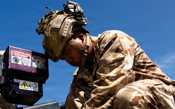 A soldier in desert camouflage uniform and tactical helmet is crouched on top of a military vehicle, performing maintenance or technical work. The soldier appears to be connecting or adjusting cables/wiring near a piece of equipment marked with danger warning labels. The image is taken from a low angle against a clear blue sky.